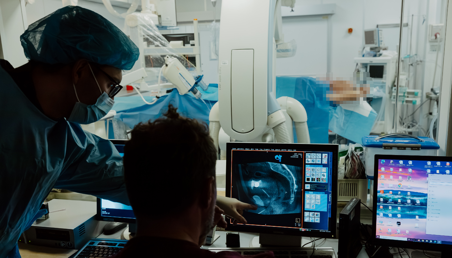 Dwie osoby w maseczkach i fartuchach patrzą na ekran z obrazem wnętrza ciała, w tle widać pacjenta przykrytego niebieskim materiałem i sprzęt medyczny.